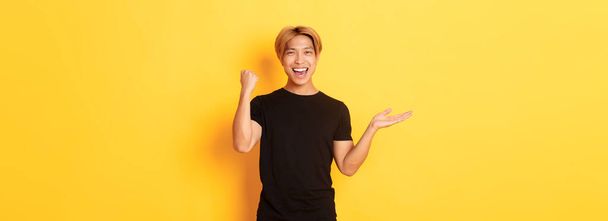 Zufrieden und glücklich attraktive koreanische Kerl lächelnd, Faust Pumpe mit Freude, hält etwas auf der Hand, stehend gelben Hintergrund. - Foto, Bild