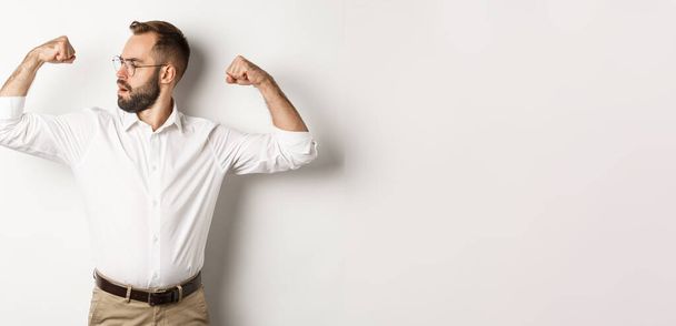 Homme d'affaires prospère flex biceps, montrant des muscles et regardant confiant, se sentant fort, debout sur fond blanc. - Photo, image