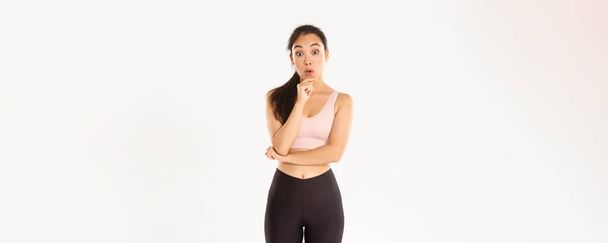 Sport, wellbeing and active lifestyle concept. Thoughtful slim and fit asian fitness girl picking gym, thinking or making decision, looking amused and wondered at camera, standing in activewear. - Photo, Image