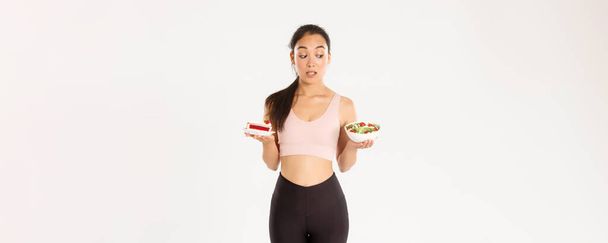 Aktives Lebens-, Fitness- und Wohlfühlkonzept. Porträt eines unentschlossenen und verführerischen süßen asiatischen Mädchens, das versucht, der Versuchung zu widerstehen, leckeren Kuchen zu halten, Diät zu halten und sich gesunden Salat anzusehen. - Foto, Bild