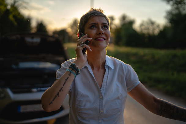 une femme mûre debout sur la route dans la soirée coucher de soleil par le véhicule cassé voiture voiture moteur défectueux capot ouvert faire un appel téléphonique pour aider assistance routière remorquage concept de service - Photo, image