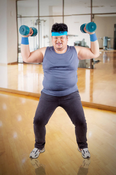 Full length of happy fat man exercising his biceps with two dumbbells while standing in the gym center - Foto, Imagen