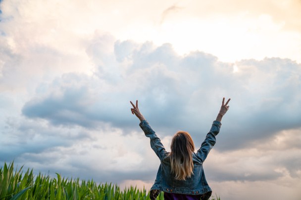 Vue arrière d'une jeune femme vêtue d'une veste en jean debout sous le ciel nuageux du soir, les deux bras levés en l'air, faisant un geste de paix. - Photo, image