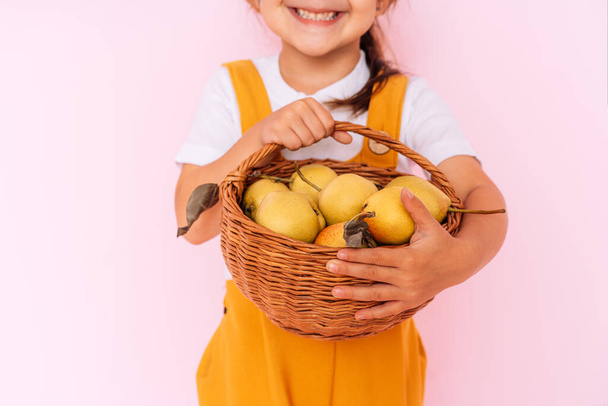 Little girl in orange overalls holding basket of pears in her hands against pink background. - 写真・画像
