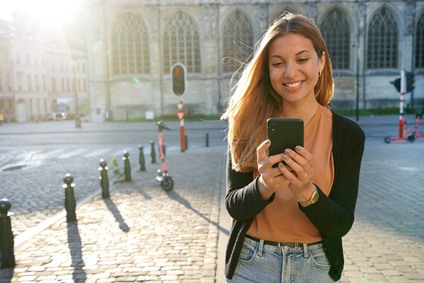 Attractive woman walking using mobile phone on urban background at sunset - Photo, Image