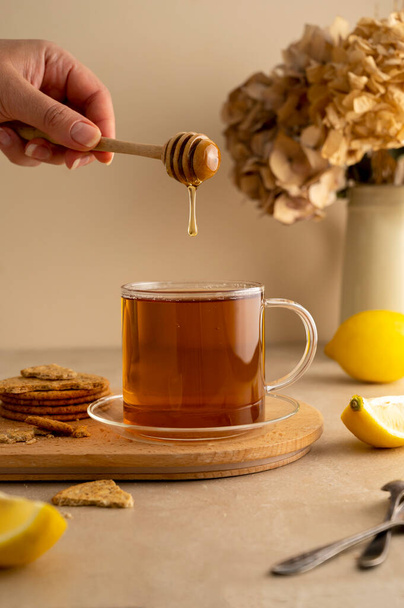 Наливаю мёд в чай. Стеклянная чашка черного чая с лимоном, теплый нейтральный фон. - Фото, изображение