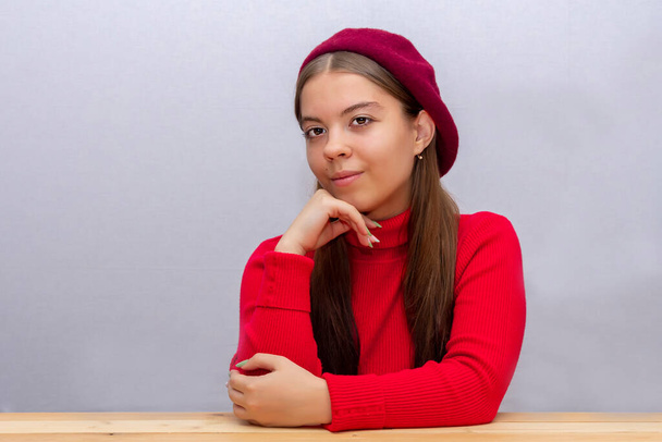 portrait of a young girl in a beret and a red sweater at a wooden table made of planks, leaning her head on her hand, light background. - Foto, imagen