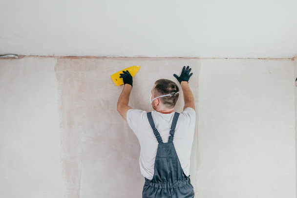 Male worker in coveralls, gloves and protective face mask applying fiberglass surface tissue on the wall using plastic spatula, preparation of surfaces for plastering or painting. Selective focus. - Foto, Imagem