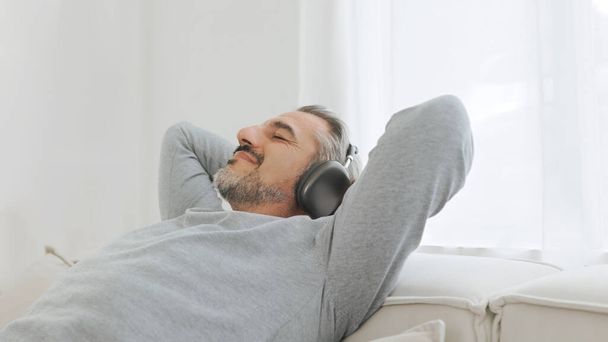 Щасливий розслаблений зрілий дорослий чоловік в навушниках слухає музику і відпочиває на зручному дивані у вітальні вдома. Старший чоловік спить на дивані
 - Фото, зображення