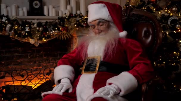 Joulupukki nukkuu istuessaan taustalla takka joulukuusi. Isoisä nukahtaa joulupukiksi pukeutuneena. Uusi vuosi joulu talvilomat juhla - Materiaali, video