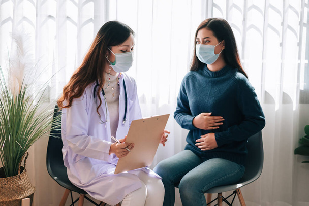 Aziatische arts bezoekt en onderzoekt jonge volwassen vrouw in het ziekenhuis met buikpijn. De arts controleert en adviseert voor de gezondheidszorg, draagt een masker ter bescherming van covid-19 pandemie. - Foto, afbeelding