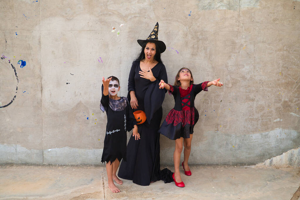 Happy Halloween. Young beautiful woman, a boy and a girl dressed up for halloween party on grey background and pumpkin in hand throwing candy in the air. Trick or treat. - Zdjęcie, obraz