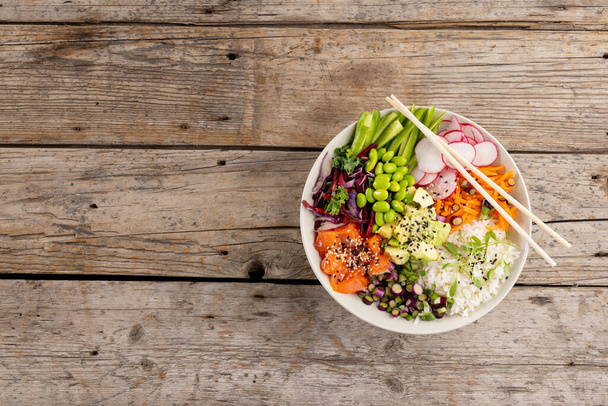 Overhead view of hawaiian poke bowl with chopsticks and copy space on wooden background. Hawaiian tasty fresh food and healthy eating. - Foto, Imagen