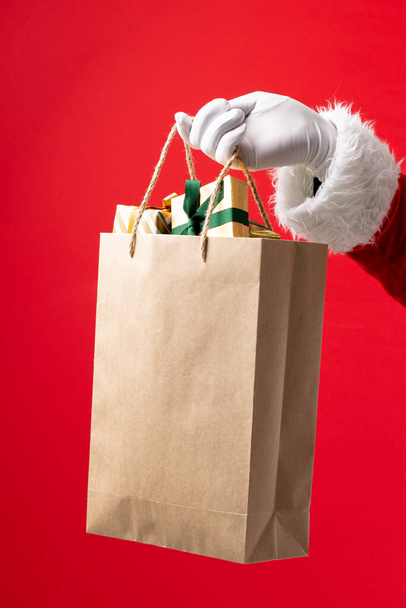 Immagine di mano di Babbo Natale che tiene il sacchetto di carta con regali di Natale su sfondo rosso. Natale, tradizione e concetto di festa. - Foto, immagini