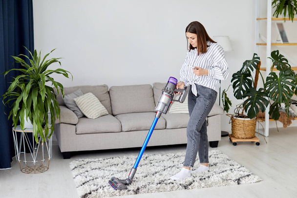 Happy cheerful young Caucasian beautiful woman vacuuming floor and using smartphone at home in living room, pretty female using vacuum cleaner, domestic concept - 写真・画像