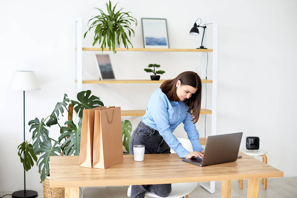 Счастливая деловая женщина, работающая в домашнем офисе стоя, глядя на свой ноутбук в окружении подарочных пакетов и упаковки - Фото, изображение
