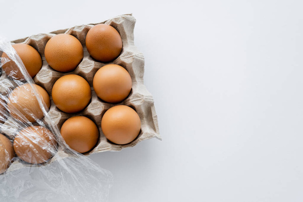 Karton tepside beyaz arka planda selofan ile kahverengi yumurtaların üst görüntüsü  - Fotoğraf, Görsel