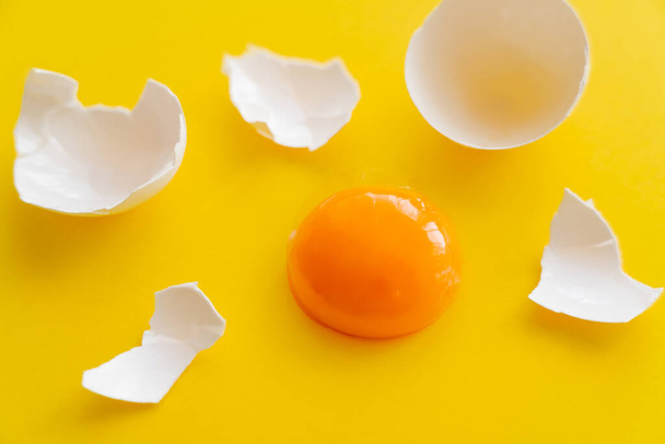 Close-up zicht op rauwe dooier in de buurt van gebarsten eierschaal op gele achtergrond  - Foto, afbeelding