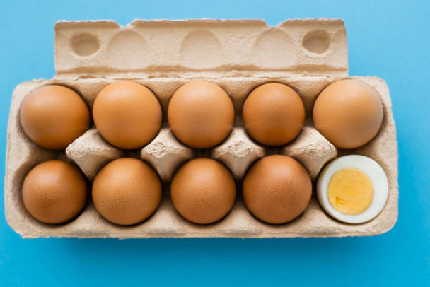 Top view of cut boiled and raw eggs in cardboard container on blue background - Fotografie, Obrázek