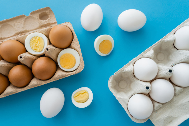 Draufsicht auf natürliche rohe und gekochte Eier in der Nähe von Pappschalen auf blauem Hintergrund - Foto, Bild