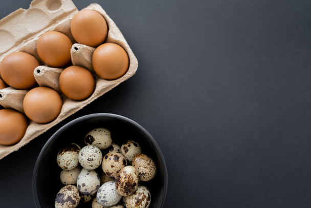 Κάτοψη του ορτυκιού και αυγών κοτόπουλου σε μπολ και δίσκο σε μαύρο φόντο  - Φωτογραφία, εικόνα