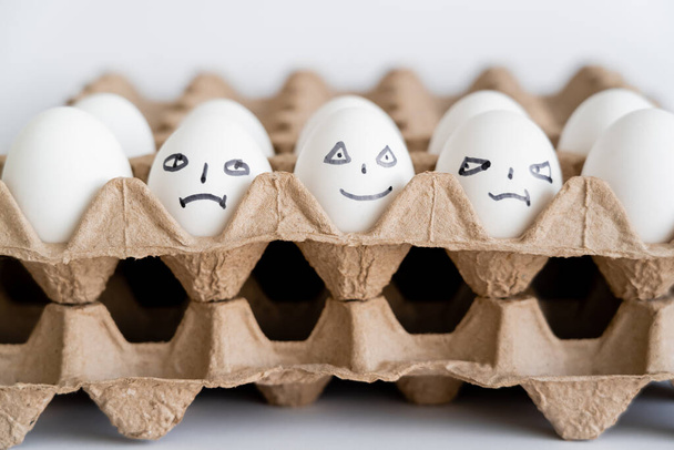 Huevos pintados con diferentes expresiones faciales en cajas de cartón sobre fondo blanco  - Foto, imagen