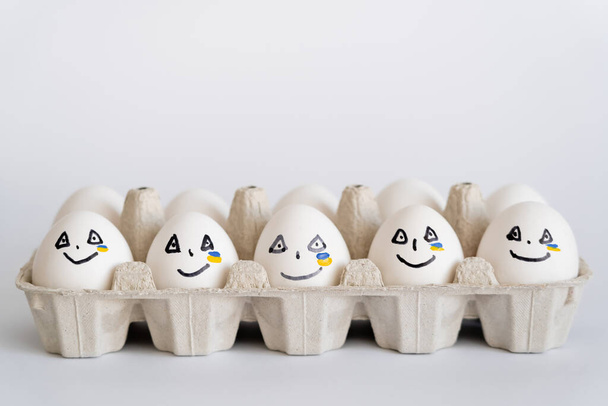 Painted eggs with facial expressions and ukrainian flag in tray on white background  - Photo, Image