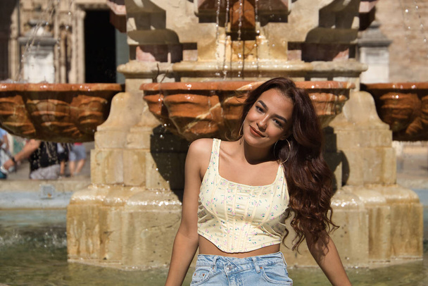 Mooie jonge vrouw met lang bruin haar en kaneelhuid zittend in een fontein, lachend, ontspannen zonnebaden op vakantie. Concept schoonheid, mode, make-up, ontspannen, vrede, rust, reizen. - Foto, afbeelding