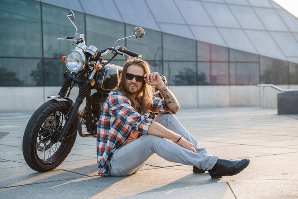 Retrato de un motociclista guay con gafas de sol sentado sobre asfalto. - Foto, Imagen