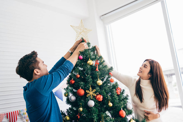 Glückliches junges Paar in Pullovern, das Spaß beim Schmücken verschiedener Kugeln am Weihnachtsbaum im heimischen Wohnzimmer hat. Das Weihnachtsfest genießen und feiern. - Foto, Bild