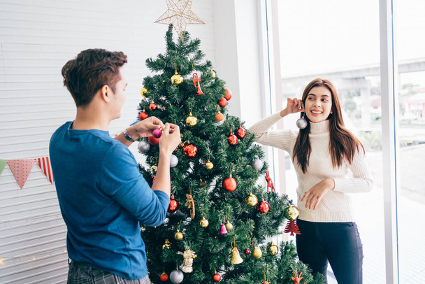 Feliz casal jovem vestindo camisolas se divertindo decorando várias bugigangas na árvore de Natal na sala de estar em casa. Desfrutando e celebrando o feriado de Natal. - Foto, Imagem