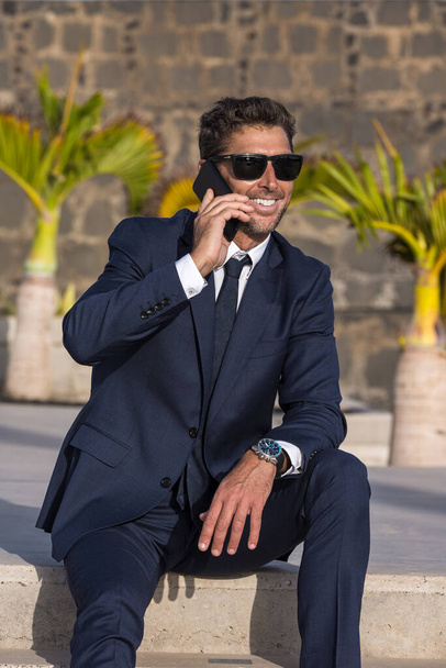 Feliz hombre de negocios con traje y gafas de sol sonriendo y mirando hacia otro lado mientras está sentado en el paso y contestando una llamada telefónica en la calle de la ciudad iluminada por el sol - Foto, Imagen