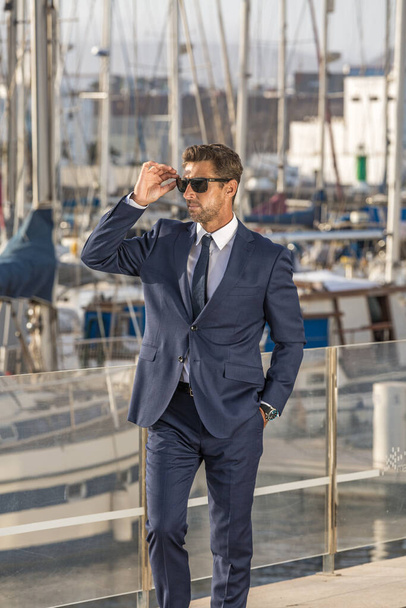 Чоловік-підприємець з рукою в кишені костюма коригує сонцезахисні окуляри і дивиться геть, стоячи на набережній проти яхт у сонячний день у місті
 - Фото, зображення