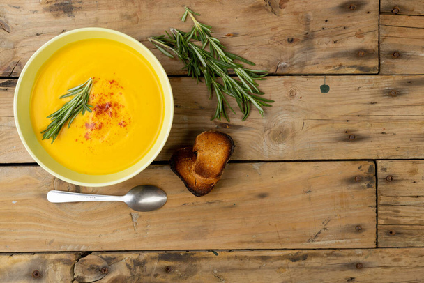Horizontal image of bowl of carrot soup with rosemary garnish, spoon and toast on wood, copy space. Tasty home cooked food and healthy eating. - Φωτογραφία, εικόνα