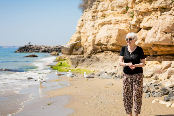 Senior woman in dark sunglasses feeds seagulls on the rocky beach. Happy life in retirement - Fotografie, Obrázek