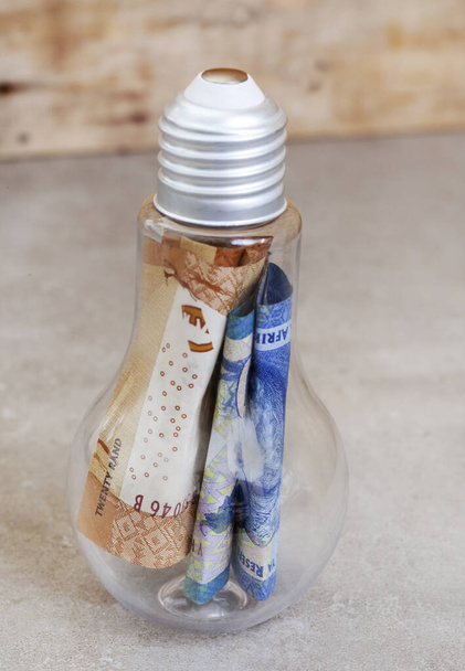 Lightbulb filled with money, South African Rand on mottled grey and selective focus - Photo, Image