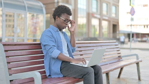 Afrikaanse man met hoofdpijn met behulp van laptop tijdens het zitten buiten op de bank - Foto, afbeelding