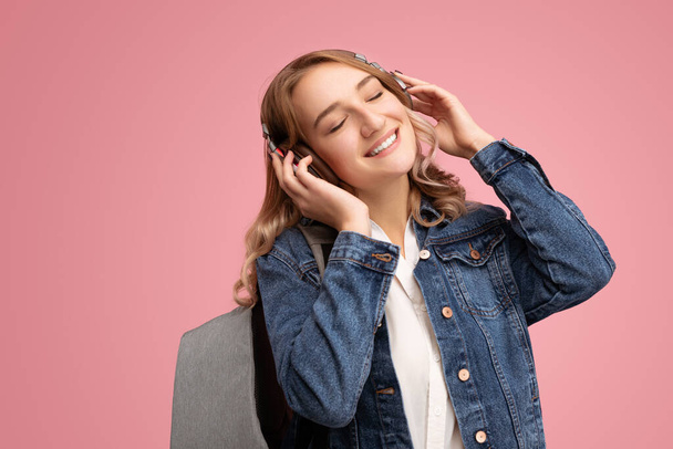 Happy young woman in denim jacket with backpack touching wireless headphones and smiling with closed eyes while enjoying good music against pink background - Fotografie, Obrázek