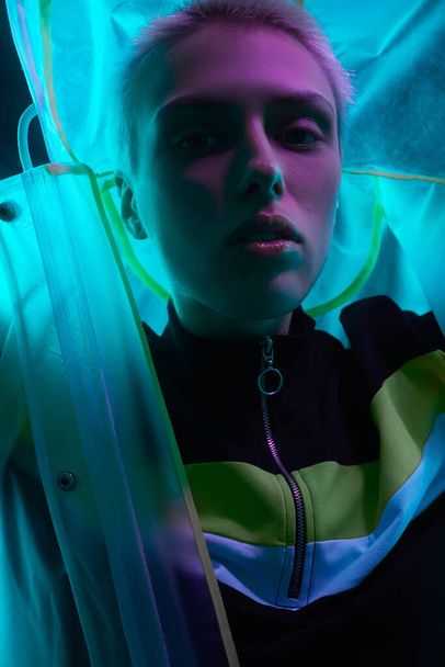 Young female model with short hair putting on hood of translucent futuristic raincoat under neon illumination - Photo, image