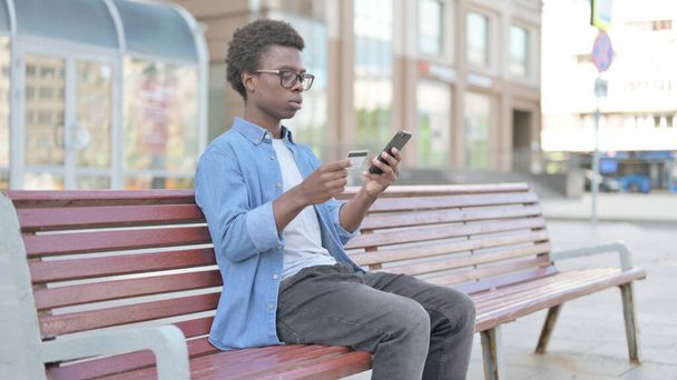 Excited African Man Shopping Online via Smartphone while Sitting Outdoor on Bench - Foto, Imagen