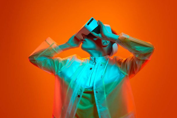 Young female gamer in futuristic clothes touching VR goggles and looking away while exploring cyberspace under turquoise neon light against orange background - Photo, image