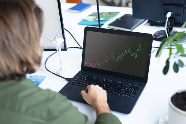 Caucasian businesswoman sitting at desk using laptop with graph and statistical data on screen. business communication and digital interface technology digital composite image. - 写真・画像
