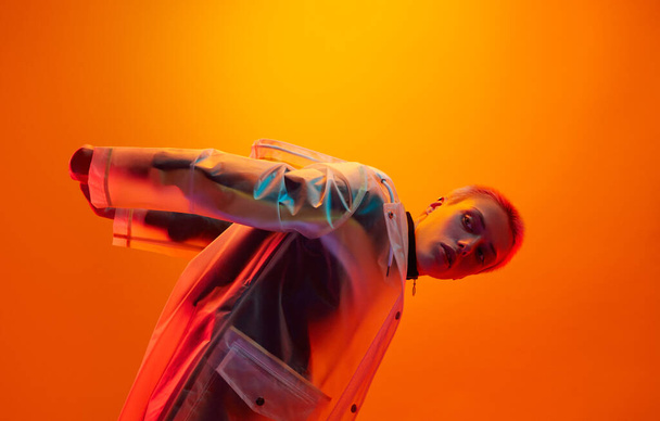 Side view of young woman in translucent futuristic raincoat holding hands behind back and looking away, while bending forward under neon illumination against orange background - Photo, image