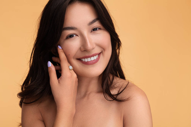 Happy Kazakh female with black hair and bare shoulders looking at camera with smile and touching cheek during skin care routine against brown background - Φωτογραφία, εικόνα