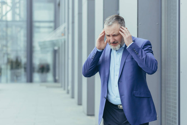 Nervous breakdown, exhaustion. Senior man businessman stands on the street near the office center and holds his head with both hands. Winced in pain, experiences nervous tension at work, needs breaks. - Fotoğraf, Görsel