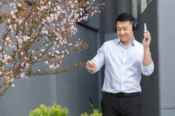 Happy young handsome male Asian student in white shirt holding phone and listening to music in headphones and dancing on campus near beautiful pink magnolia tree. - Zdjęcie, obraz