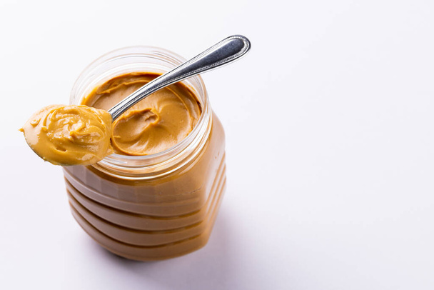 Image of spoon and jar with peanut butter on white surface. Food, breakfast, nutrition and american cuisine concept. - Foto, Imagen