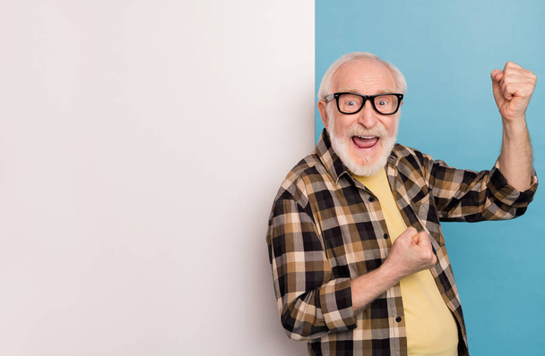 Foto van schattige gelukkige man gepensioneerde gekleed geruite shirt brillen stijgende vuisten billboard lege ruimte geïsoleerde blauwe kleur achtergrond. - Foto, afbeelding