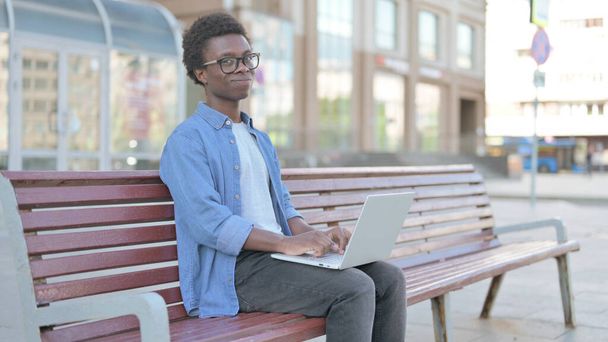 Відкидаючи африканця у запереченні, використовуючи лаптоп сидячи надворі на лаві - Фото, зображення