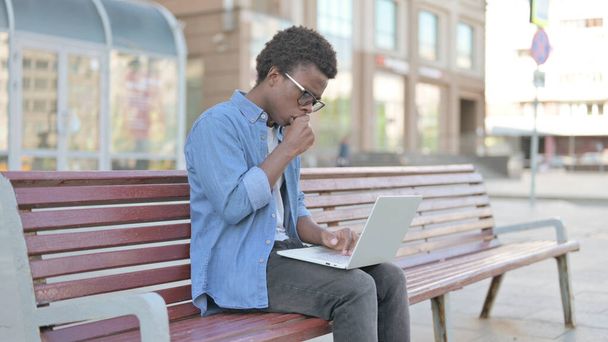 Hoesten Afrikaanse man met behulp van laptop tijdens het zitten buiten op de bank - Foto, afbeelding